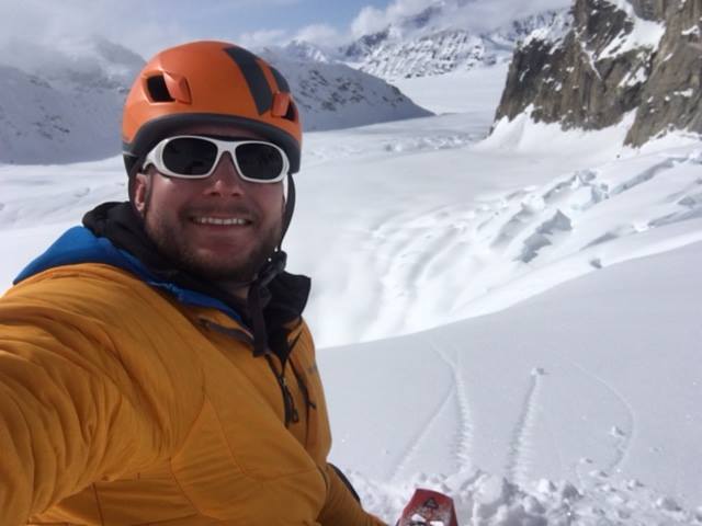 A man wearing outdoor gear and sunglasses, smiling as he take a selfie in from of snow-covered mountains